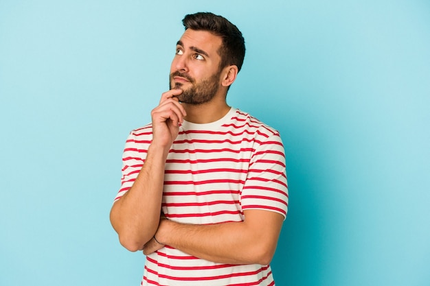 Young caucasian man isolated on blue wall thinking and looking up, being reflective, contemplating, having a fantasy