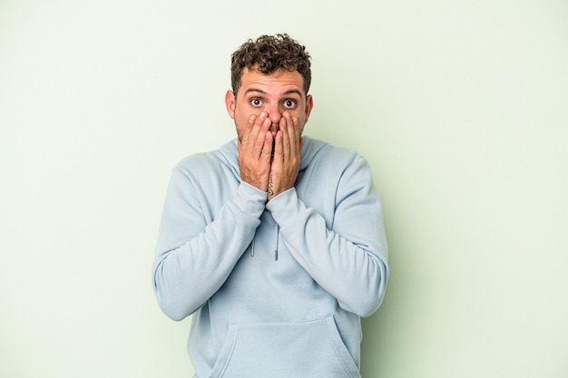 Young caucasian man isolated on green background shocked, covering mouth with hands, anxious to discover something new.