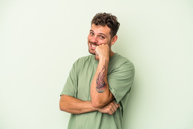 Young caucasian man isolated on green background smiling happy and confident, touching chin with hand.