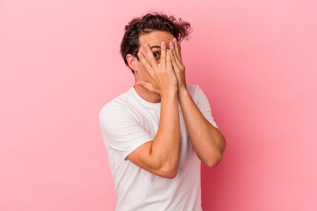 Young caucasian man isolated on pink background blink through fingers frightened and nervous.