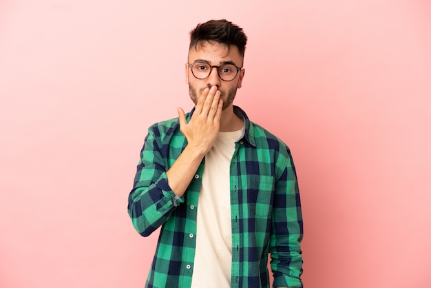 Young caucasian man isolated on pink background covering mouth with hand