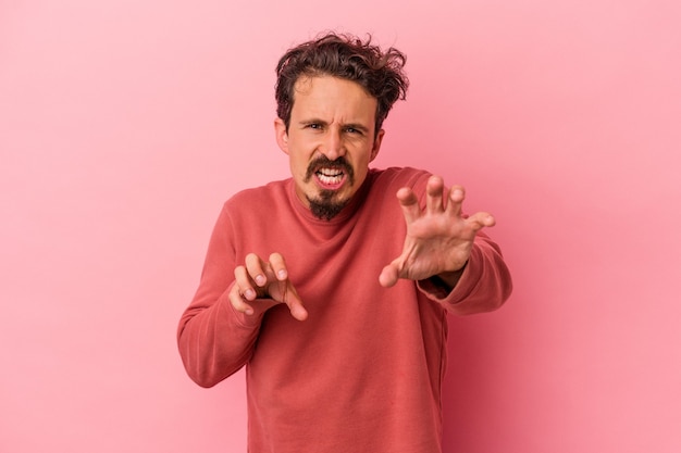 Young caucasian man isolated on pink background showing claws imitating a cat, aggressive gesture.