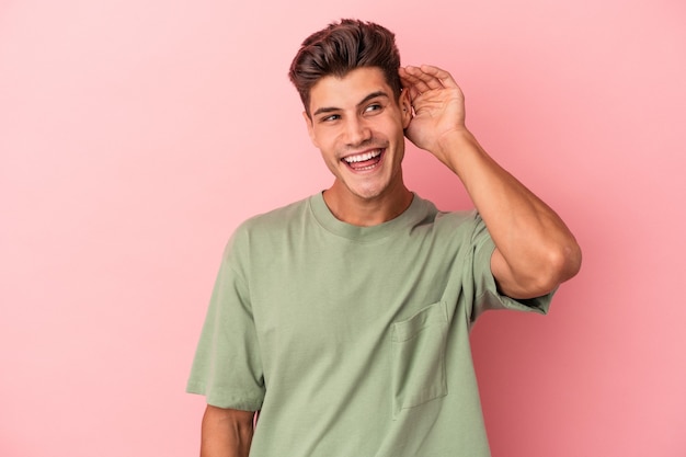 Young caucasian man isolated on pink background trying to listening a gossip.