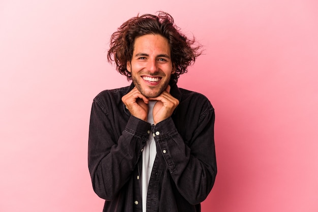 Young caucasian man isolated on pink bakcground keeps hands under chin, is looking happily aside.