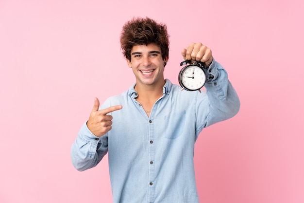 Young caucasian man over isolated pink wall holding vintage alarm clock