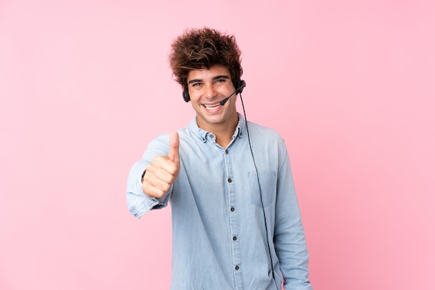 Young caucasian man over isolated pink wall working with headset with thumb up