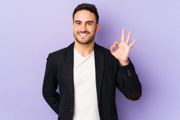 Young caucasian man isolated on purple space winks an eye and holds an okay gesture with hand.