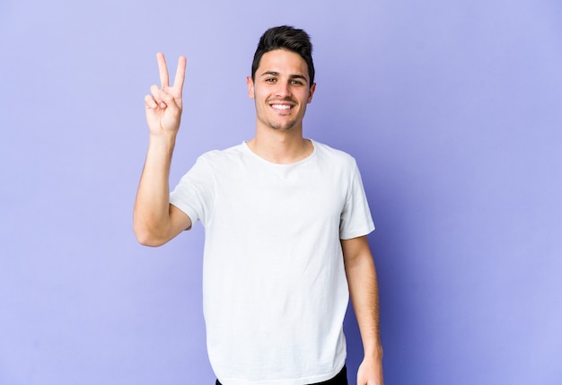 Young caucasian man isolated on purple wall showing victory sign and smiling broadly.