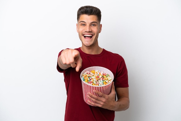 Young caucasian man isolated on white background holding a big bucket of popcorns while pointing front