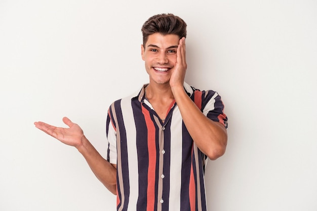 Young caucasian man isolated on white background holds copy space on a palm, keep hand over cheek. Amazed and delighted.