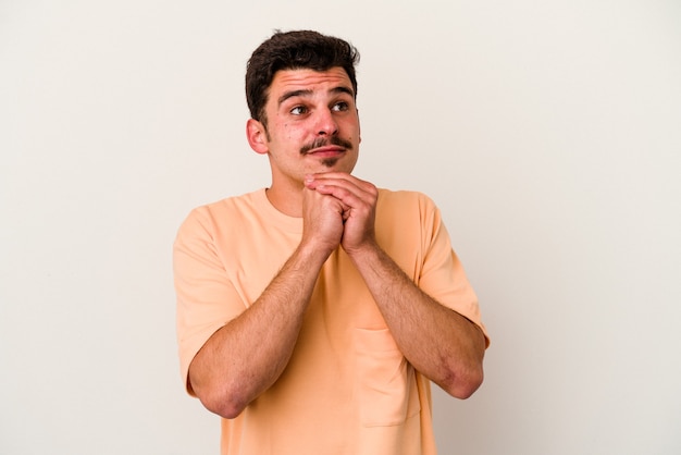 Young caucasian man isolated on white background keeps hands under chin, is looking happily aside.