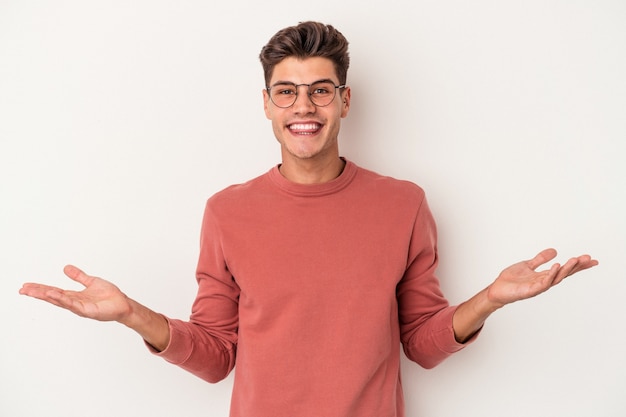 Young caucasian man isolated on white background makes scale with arms, feels happy and confident.