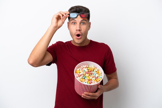 Young caucasian man isolated on white background surprised with 3d glasses and holding a big bucket of popcorns