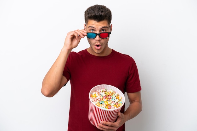 Young caucasian man isolated on white background surprised with 3d glasses and holding a big bucket of popcorns