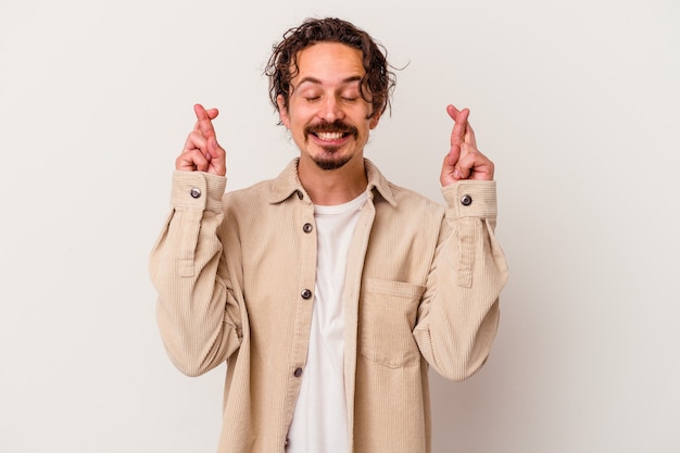 Young caucasian man isolated on white wall crossing fingers for having luck