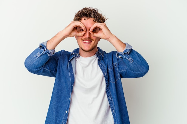 Young caucasian man isolated on white wall showing okay sign over eyes