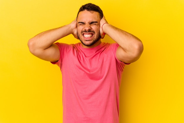 Young caucasian man isolated on yellow background covering ears with hands trying not to hear too loud sound.