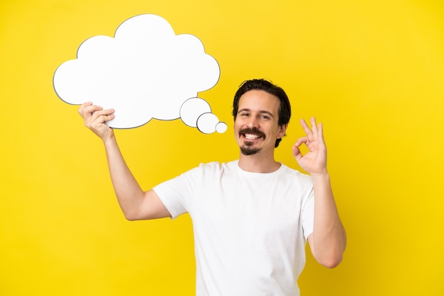 Young caucasian man isolated on yellow background holding a thinking speech bubble and doing OK sign