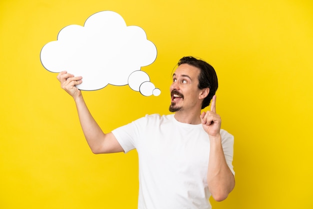 Young caucasian man isolated on yellow background holding a thinking speech bubble with surprised expression