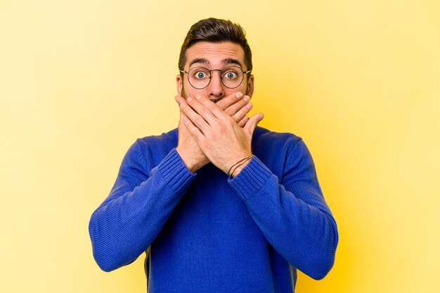 Young caucasian man isolated on yellow background shocked covering mouth with hands