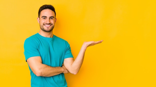 Young caucasian man isolated on yellow showing a copy space on palm and holding another hand on waist.