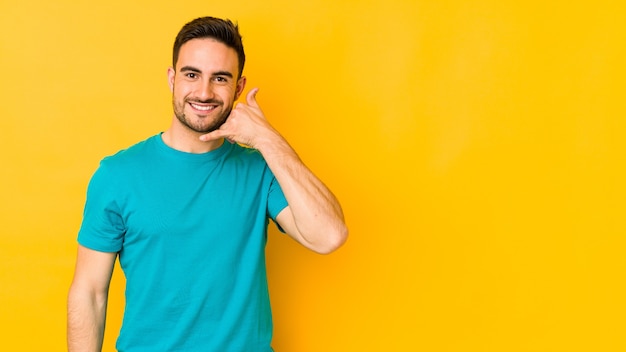 Young caucasian man isolated on yellow wall showing a mobile phone call gesture with fingers.