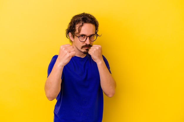 Young caucasian man isolated on yellow wall throwing a punch, anger, fighting due to an argument, boxing