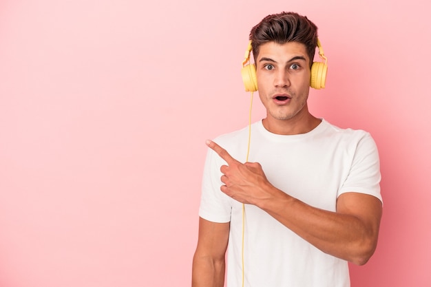 Young caucasian man listening to music isolated on pink background pointing to the side