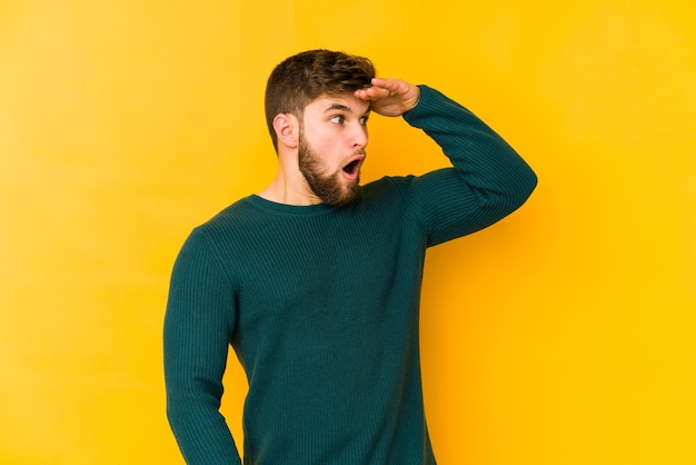 Young caucasian man looking far away keeping hand on forehead.