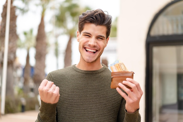 Young caucasian man at outdoors holding wallet with money and celebrating a victory