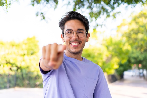 Young caucasian man at outdoors in a park points finger at you with a confident expression