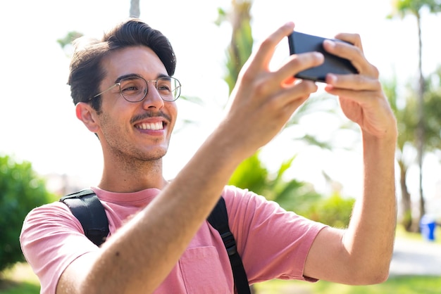 Young caucasian man at outdoors in a park using mobile phone and doing a selfie