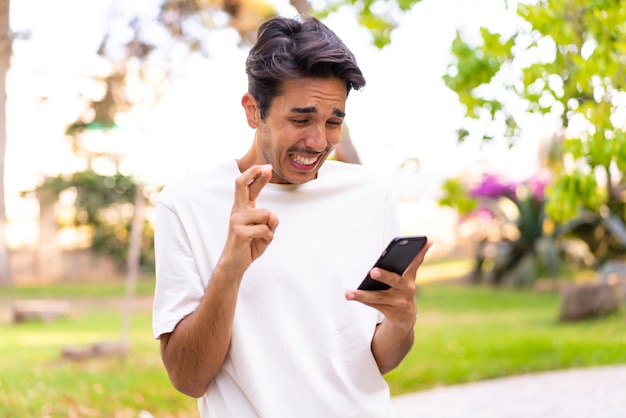 Young caucasian man in a park using mobile phone with fingers crossing