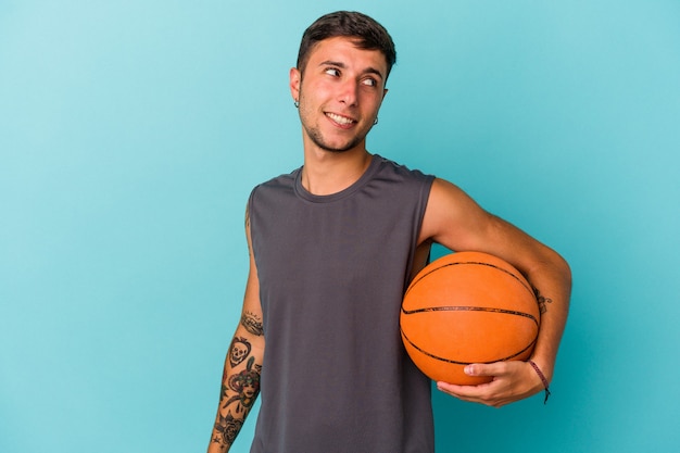 Young caucasian man playing basketball isolated on blue background  looks aside smiling, cheerful and pleasant.