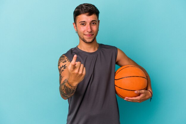 Young caucasian man playing basketball isolated on blue background  pointing with finger at you as if inviting come closer.