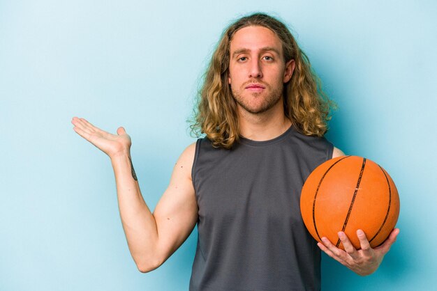 Photo young caucasian man playing basketball isolated on blue background showing a copy space on a palm and holding another hand on waist