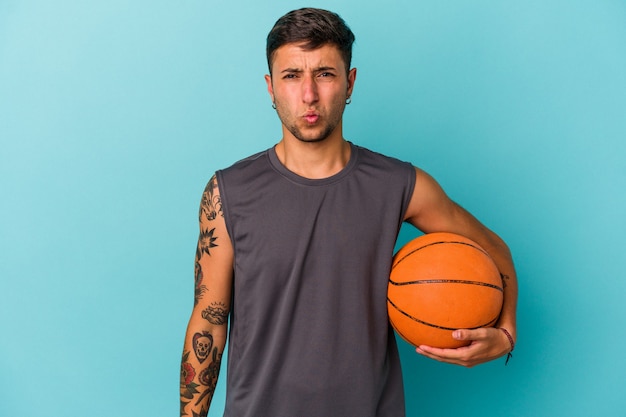 Young caucasian man playing basketball isolated on blue background  shrugs shoulders and open eyes confused.