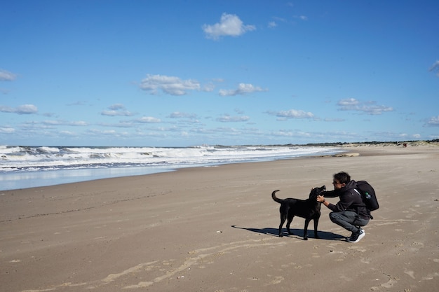 Young Caucasian man playing on the beach with black dog. On sand under light blue sky with clouds.