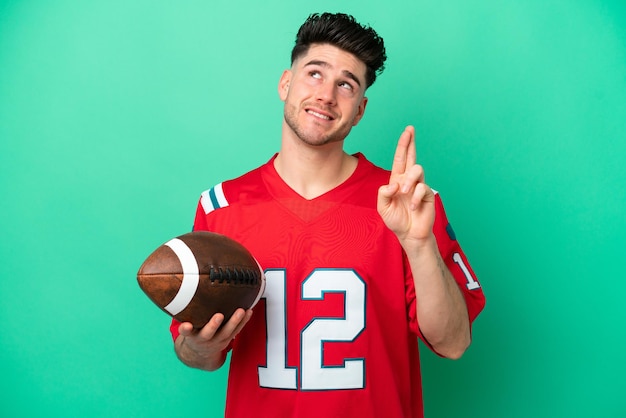 Young caucasian man playing rugby isolated on green background with fingers crossing and wishing the best