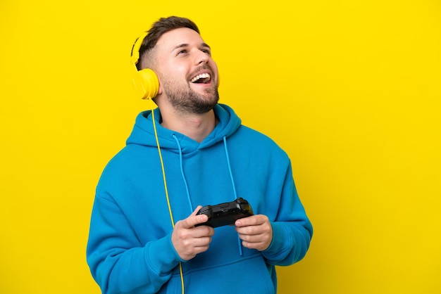 Young caucasian man playing with a video game controller isolated on yellow background laughing