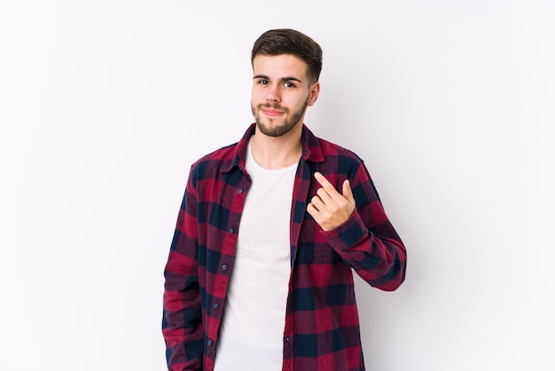 Young caucasian man posing in a white wall isolated pointing with finger at you as if inviting come closer.