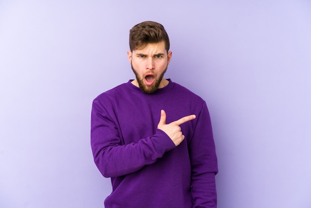 Young caucasian man on purple wall pointing to the side