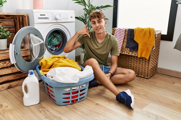 Young caucasian man putting dirty laundry into washing machine smiling doing phone gesture with hand and fingers like talking on the telephone. communicating concepts.