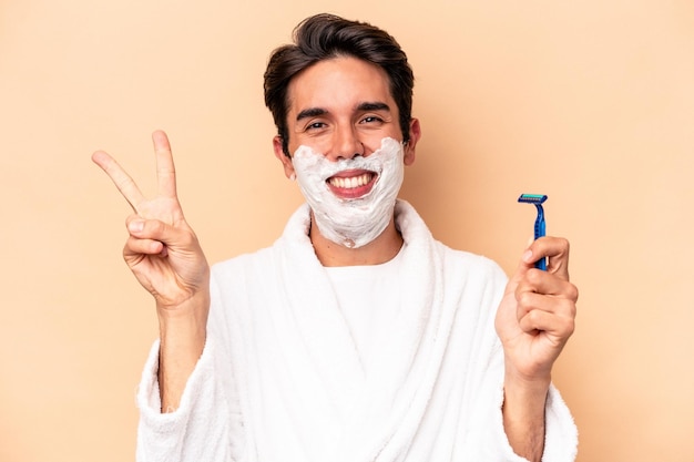 Young caucasian man shaving his beard isolated on beige background joyful and carefree showing a peace symbol with fingers.