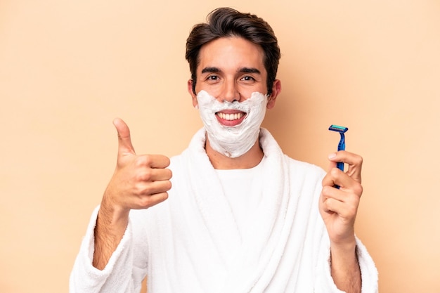 Young caucasian man shaving his beard isolated on beige background smiling and raising thumb up