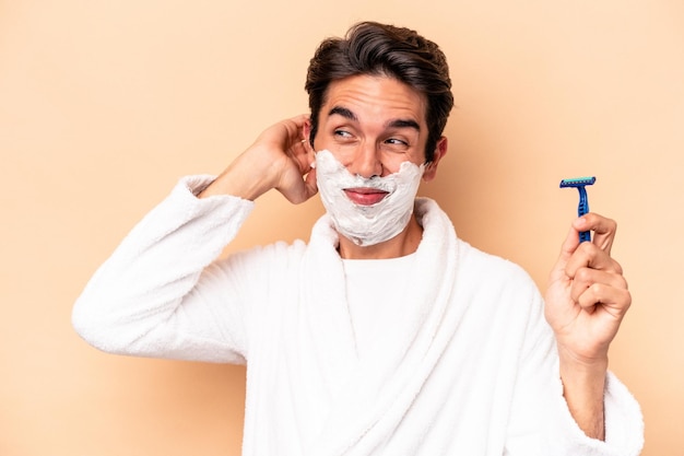 Young caucasian man shaving his beard isolated on beige background touching back of head, thinking and making a choice.