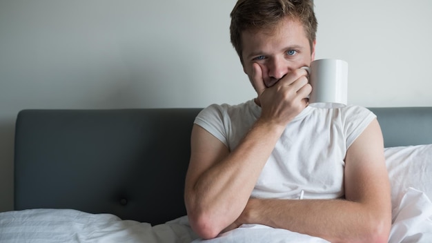 Photo young caucasian man sitting on bed in bedroom with cup of water