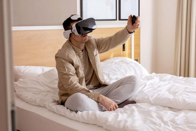 Young caucasian man sitting on bed at home with VR headset and playing interactive video game exploring virtual reality Man wearing VR glasses Future gadgets technology education study online