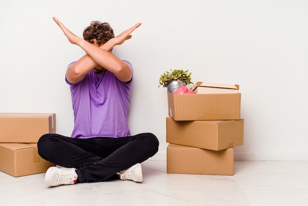 Photo young caucasian man sitting on the floor ready for moving isolated on white background keeping two arms crossed, denial concept.