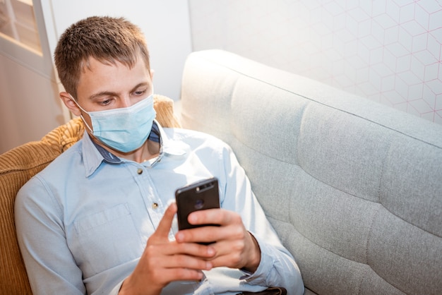 Young caucasian man sitting on a sofa in casual clothes wearing a protective medical mask with a pho...